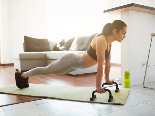 Young woman doing sport workout in room during quarantine. Picture of strong fitness model stand in plank position using push up stands hand bar. Exercising in room alone.